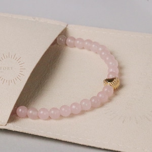 May include: A light pink beaded bracelet with a gold heart charm, resting on a cream-colored pouch. The bracelet is made of small, round beads. The pouch has the text "OHMYGEM STORY HANDMADE JEWELLERY" printed on it.