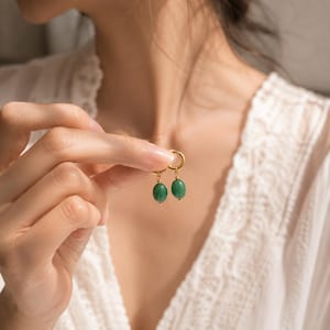 May include: A pair of gold hoop earrings featuring small, oval-shaped green stone charms. Each earring consists of a thin, circular gold hoop with a single, smooth, deep green bead suspended from the bottom, creating a delicate and simple design.