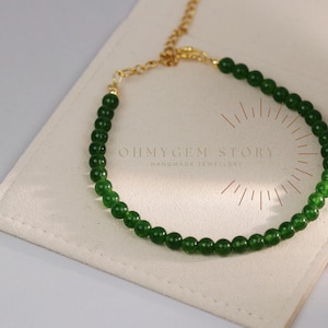 May include: A bracelet with small, round, dark green beads and a gold-coloured chain. The bracelet is displayed on a cream-coloured pouch with the text "OHMYGEM STORY HANDMADE JEWELLERY".