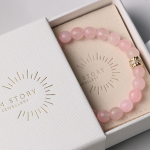 May include: A pink rose quartz bead bracelet with a gold accent bead is displayed in an open white box. The box contains a beige fabric. The box has the text "OHMYGEM STORY HANDMADE JEWELLERY" printed in gold.