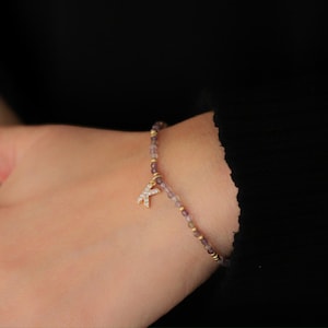 Könnte beinhalten: Ein zartes Perlenarmband mit kleinen lila und goldenen Perlen. Ein goldfarbener Anhänger in Form des Buchstabens "K" ist mit kleinen, klaren Kristallen verziert. Das Armband wird an einem Handgelenk vor schwarzem Hintergrund getragen.