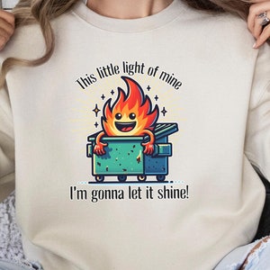 May include: Beige sweatshirt featuring a cartoon flame character in a dumpster with the text "This little light of mine, I'm gonna let it shine!" The flame is orange and yellow, and the dumpster is teal. The sweatshirt is a neutral color.