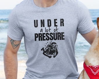 May include: A man wearing a grey t-shirt with the text "UNDER a lot of PRESSURE" and a scuba diver graphic. He is sitting on the beach with a dog wearing a red bandana.