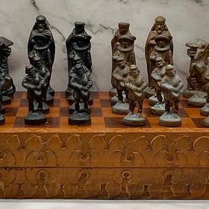 May include: A wooden chess set featuring a rectangular board with carved decorative edges and metal hinges. The set includes a collection of black and metallic-toned chess pieces arranged on the board, depicting various figures in historical attire.