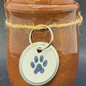 May include: A brown ceramic pet urn with a lid, tied with twine. A silver tag with a blue paw print hangs from the urn. The urn has a glossy finish and a classic shape, suitable for pet memorials.