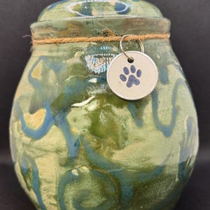 May include: A ceramic pet urn with a lid, glazed in shades of green and blue with a swirling pattern. A small, round tag with a blue paw print hangs from a twine tied around the urn. The urn is designed to hold the ashes of a beloved pet.