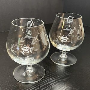 May include: Two clear brandy snifters with etched floral designs. The glasses have a rounded bowl shape, a stem, and a circular base. The etched design features leaves and circular patterns. The glasses are on a dark surface.