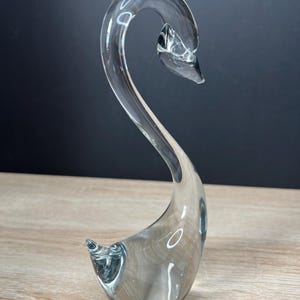 May include: A clear glass swan figurine with a curved neck and body. The swan is displayed on a light wood surface against a dark background. The elegant sculpture is a decorative item.