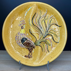 May include: A decorative, mustard-yellow ceramic plate featuring a raised relief of a rooster. The rooster has a detailed design with brown, black, yellow, and red accents. The plate is round and has a textured surface, with a dark background.