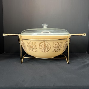 May include: A cream-colored ceramic casserole dish featuring a gold snowflake-like pattern, topped with a clear glass lid. The dish is held within a gold-toned metal wire stand that includes two horizontal side handles for easy carrying.