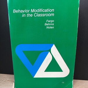 May include: A green book titled "Behavior Modification in the Classroom" with the authors Fargo, Behrns, and Nolen. The cover features a blue and white geometric design. The book is standing upright on a light-colored surface.
