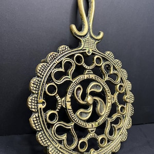 May include: A circular brass trivet featuring an intricate openwork design with heart-shaped cutouts, a central four-lobed motif, and a scalloped outer edge. The piece includes an integrated handle at the top for hanging, with a dark patina.