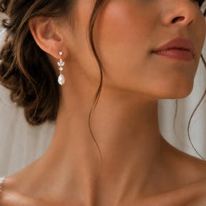 May include: A gold-toned drop earring featuring a small round clear stone at the top, followed by a marquise-shaped clear stone, a small round clear stone, and a white teardrop-shaped pearl dangling at the bottom of the vertical arrangement.