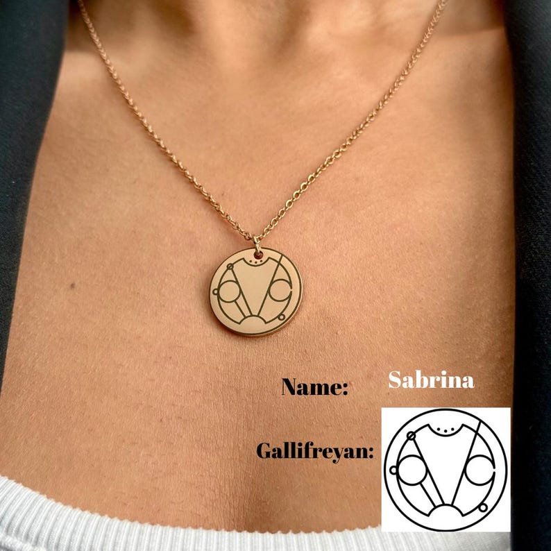 May include: A gold necklace with a round pendant featuring a Gallifreyan symbol. The pendant is engraved with a geometric design. The necklace is worn by a person, and the text "Name: Sabrina" and "Gallifreyan:" is visible below the image.