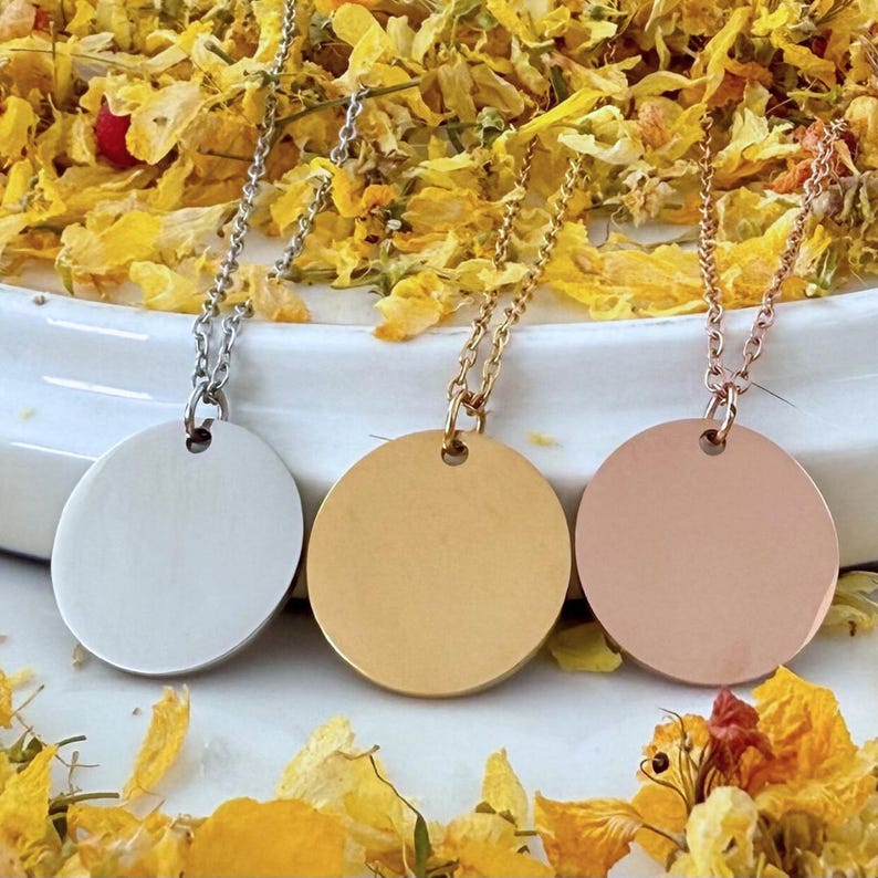 May include: Three necklaces with round, blank, silver, gold, and rose gold pendants. The necklaces are on a white surface with yellow flower petals.