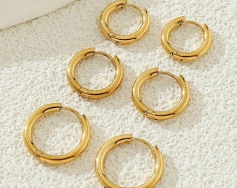 May include: Six gold hoop earrings. The earrings are circular with a hinged clasp. The earrings are made of a shiny, metallic material. The earrings are displayed on a textured, off-white surface.