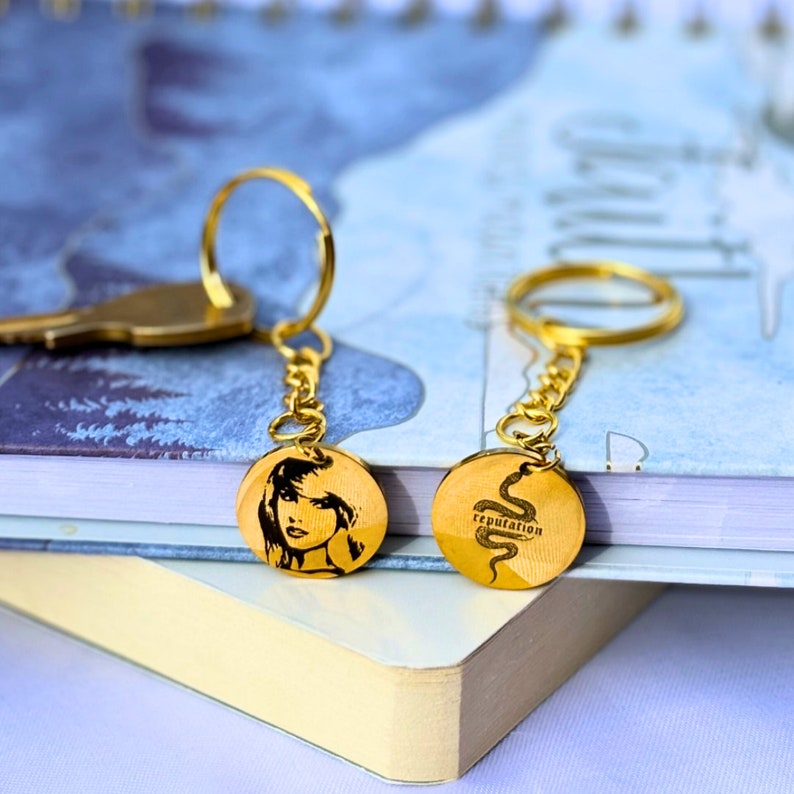 May include: Two gold keychains with round metal charms. One charm has a black and white image of a woman's face. The other charm has a black and white image of a snake with the word 'reputation' above it.