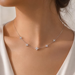 May include: A delicate silver necklace featuring five flower-shaped pendants. Each pendant is adorned with small, sparkling stones, creating a subtle yet elegant design. The necklace is shown against a neutral background, highlighting its refined aesthetic.