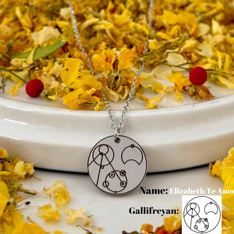 May include: A silver necklace with a round pendant featuring a Gallifreyan symbol. The pendant is on a white surface with yellow flower petals scattered around it. The text "Name: Elizabeth Te Amo" and "Gallifreyan:" is visible below the pendant.