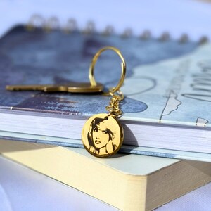May include: Gold keychain with a round charm featuring a black and white image of a woman's face.