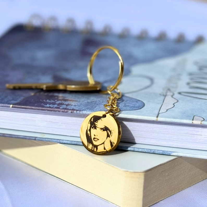 May include: Gold keychain with a round charm featuring a black and white image of a woman's face.