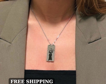 May include: A silver necklace with a pendant featuring a silhouette of a person standing in a doorway. The pendant is made of two separate pieces that are connected by a chain.