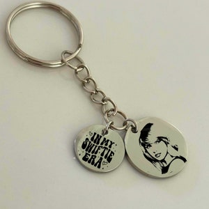 May include: Silver keychain with two round charms. One charm has a black and white image of a woman with the text "com" on the bottom. The other charm has the text "In My Swiftie Era" with hearts and stars.
