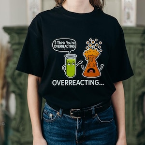 Puede incluir: Camiseta negra con un gráfico de temática científica. El diseño presenta dos vasos de dibujos animados, uno verde y tranquilo, el otro naranja y en erupción, con el texto "I Think You're Overreacting..." y "Overreacting..." debajo.
