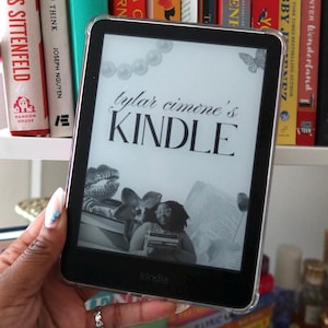 Puede incluir: Un lector de libros electrónicos Kindle negro que muestra el texto "tylar cimone's KINDLE" con una imagen de una persona leyendo. El lector de libros electrónicos está frente a una estantería llena de libros coloridos.