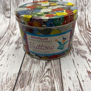 May include: A round metal tin filled with colorful buttons. The lid and sides of the tin are decorated with various buttons in shades of red, yellow, blue, green, and white. The tin has text that reads "500-PLUS Guaranteed Washable Buttons in Matched Sets."