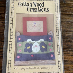 May include: A craft pattern for a pillow and a small embroidery piece. The pillow features a white bunny with yellow flowers and pink buttons. The embroidery piece is a red frame with a white bunny and clouds. The pattern is titled "Spring Buds Pillow 10" x 14" & Stitchery 5" x 7" #013"