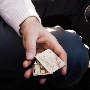 以下が含まれることがあります： ヒンジ付きの蓋が付いた、小さな長方形の銀色の金属製缶。蓋には「WILDERBRUSH」と「NATURAL SOAP」というエンボス加工の文字が刻まれています。開いた缶の中には、小さな正方形の薄茶色のテクスチャーのある石鹸キューブがいくつか入っています。