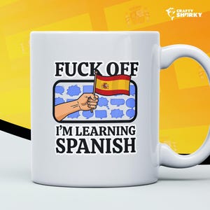 May include: White ceramic mug with a humorous graphic. The mug features the text "FUCK OFF I'M LEARNING SPANISH" in bold black letters. A hand holding a Spanish flag is also depicted. The mug has a white handle and a yellow background.
