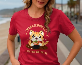 May include: Red t-shirt featuring a cartoon cat design. The cat is orange and white, holding a bowl of seafood. Text on the shirt reads "I'M ON A SEAFOOD DIET. I SEE FOOD AND I EAT IT."