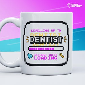 May include: White ceramic mug featuring a pixelated design that reads "Levelling up to Dentist" with a nearly full progress bar. The progress bar is pink and the text is yellow. A blue diamond and a gold key are positioned on either side of the progress bar. The text "Please wait loading" is displayed beneath the progress bar.