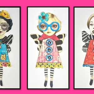 May include: Three paper dolls with wings, each wearing a different outfit. The doll on the left is wearing a blue dress with yellow and black accents, the middle doll is wearing a pink dress with blue and white accents, and the doll on the right is wearing a red dress with black and white accents.