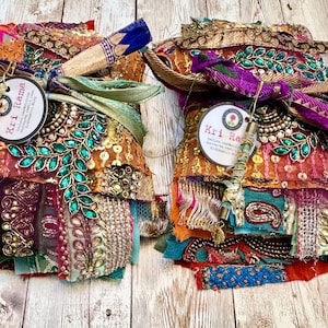 Puede incluir: Dos paquetes de retazos de tela variados con colores vibrantes como naranja, morado y turquesa. Los textiles están adornados con bordados metálicos dorados, lentejuelas y motivos de hojas con piedras preciosas verdes. Cada paquete incluye una etiqueta redonda.