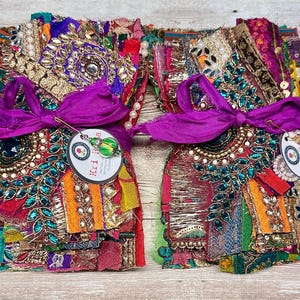 May include: Two bundles of assorted fabric scraps featuring vibrant colors, metallic gold embroidery, green faux gemstones, and white pearls. Each bundle is tied with a purple silk ribbon and includes a round tag with the text Kri Pa.