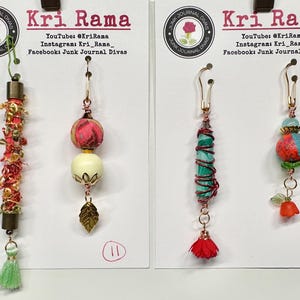May include: Four decorative charms attached to white display cards labeled Kri Rama. The charms feature various beads, fabric textures, metal wire, and small tassels in shades of red, green, blue, and gold. Each charm includes a metal hook for hanging.