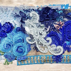 May include: A collection of blue and white lace appliques featuring floral motifs, intricate embroidery, and beadwork. The assortment includes layered fabric pieces in shades of navy, royal blue, and light blue, accented with metallic gold trim.