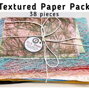 May include: A stack of 38 pieces of textured paper in various colors, including pink, blue, yellow, and green, tied with twine. The paper pack has a tag that reads "Kri Rama". The words "Textured Paper Pack" and "38 pieces" are printed at the top.