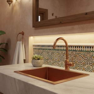 Könnte beinhalten: Ein kupferfarbenes rechteckiges Waschbecken mit passender Armatur ist in eine Marmorarbeitsplatte eingelassen. Das Badezimmer verfügt über eine dekorative Fliesenrückwand, ein Handtuch und eine Topfpflanze. Eine dekorative Wandleuchte und ein großer Spiegel vervollständigen die Szene.