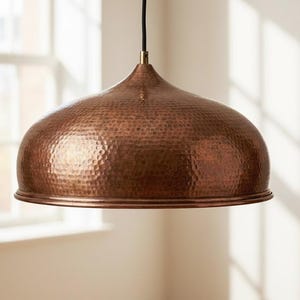 May include: A copper-colored pendant light fixture with a hammered texture. The light has a dome shape and is suspended from a black cord. The fixture is hanging in front of a window.