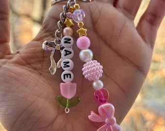 May include: A pink and white beaded keychain with a charm that says "NAME" and a pink bow. The keychain also features a pink flower charm and a green leaf charm.