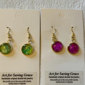 May include: Two pairs of handmade alcohol ink earrings. One pair is green and gold, the other is pink and gold. The earrings are displayed on white cards with the text "Art for Saving Grace" and information about the non-profit charity.