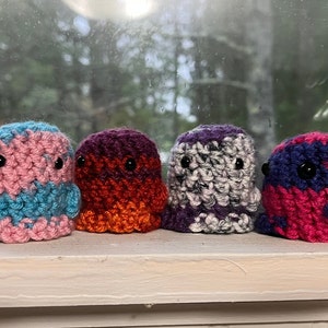 May include: Four crocheted ghost-shaped plushies in different colour combinations: pink and blue, orange and purple, grey and purple, and pink and purple.