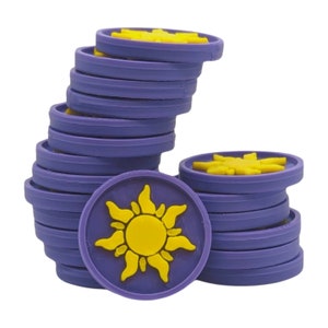 Puede incluir: Un conjunto de adornos circulares morados y amarillos. Cada adorno presenta un dise&ntilde;o de sol amarillo sobre un fondo morado. Los adornos est&aacute;n apilados en dos montones separados, algunos planos.