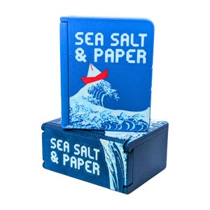 Può includere: Due scatole blu sovrapposte con la scritta "SEA SALT & PAPER". La scatola superiore presenta un motivo a onde con una barca a vela rossa. La scatola inferiore ha lo stesso testo.