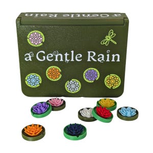 Op de afbeelding: Een olijfgroene doos met de woorden "a Gentle Rain" en bloemmotieven. De doos heeft een scharnierend deksel en is omgeven door verschillende kleine, kleurrijke, bloemvormige objecten in verschillende kleuren, waaronder paars, roze en geel.