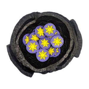 Puede incluir: Un cuenco negro texturizado lleno de fichas moradas y amarillas con forma de sol. Las fichas tienen un dise&ntilde;o de sol amarillo sobre un fondo morado. El cuenco tiene un aspecto rugoso y en capas.